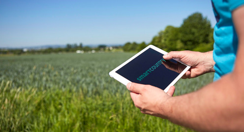 Auf dem Foto sind zwei Hände zu sehen, die ein Tablet halben. Im Hintergrund ist ein grünes Feld zu sehen.