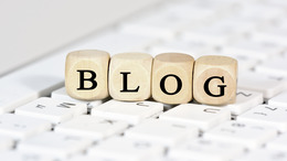 Auf einer Tastatur stehen kleine Holzwürfel mit Buchstaben, die das Wort "Blog" ergeben